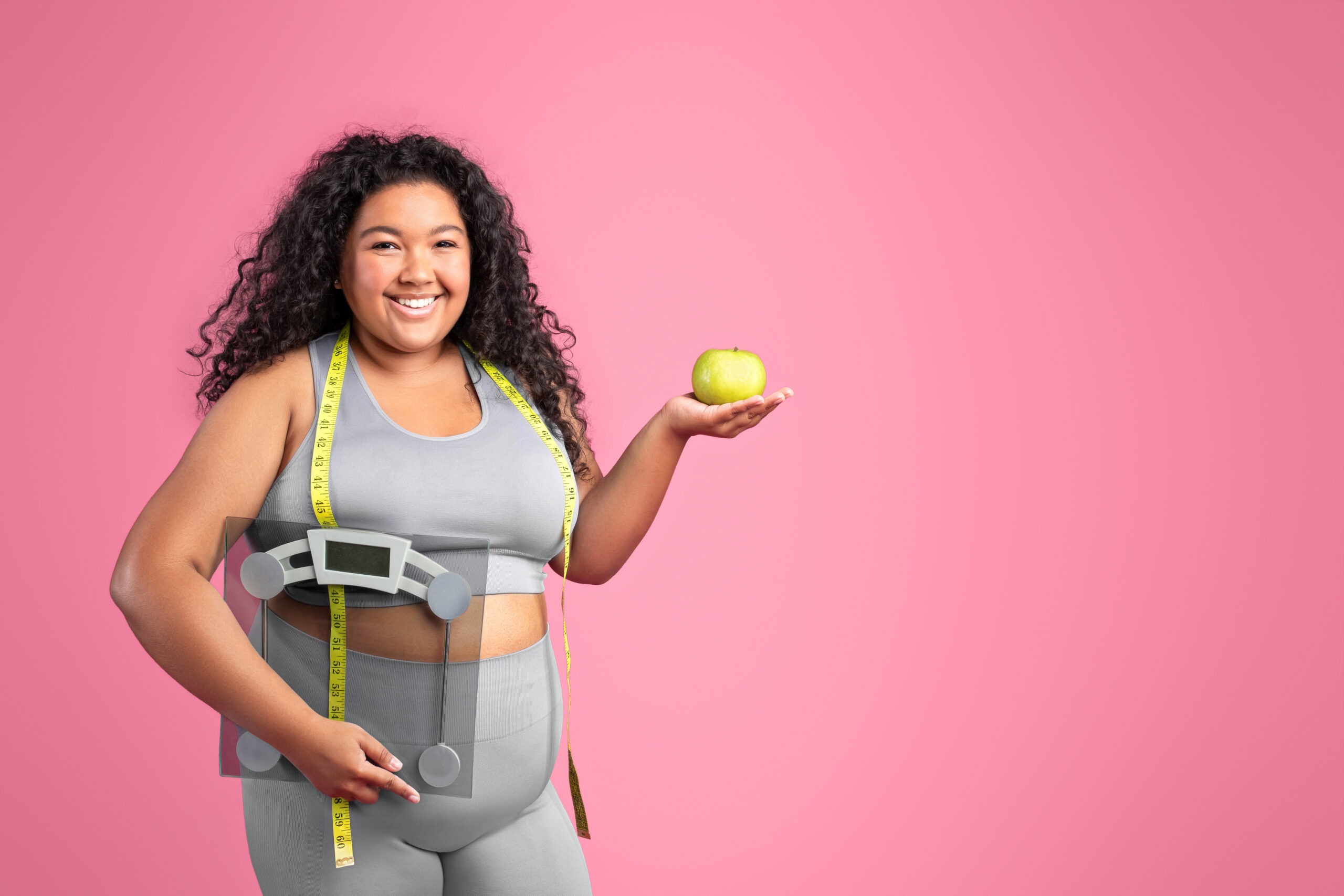 Glad black body positive woman in sportswear holding apple and scales, enjoying diet result, standing over pink studio background, copy space. Fit lifestyle, body care and healthy food