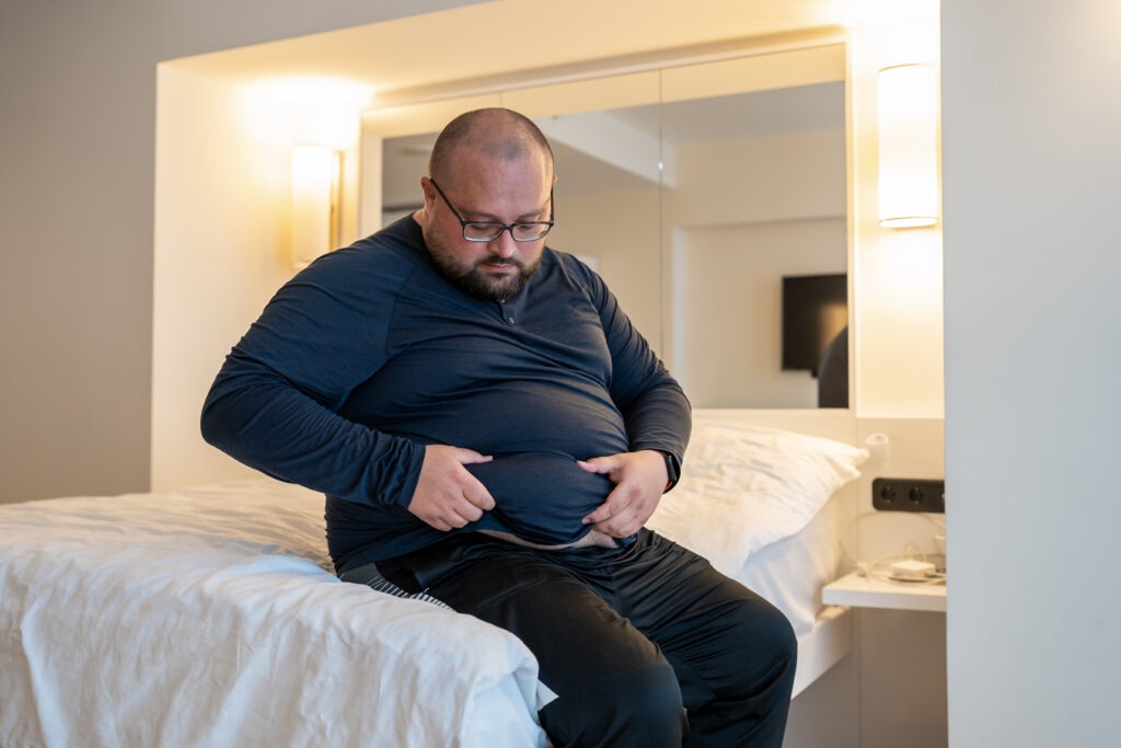 Man suffering from extra weight touching abdomen sitting on bed in bedroom. Overweight bearded balded middle aged male inspecting body to change life habits. Unhealthy body, overeating concept.