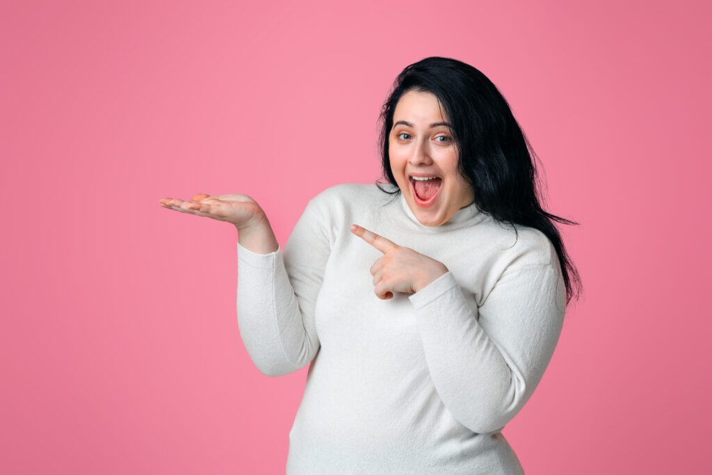 Overjoyed plump woman pointing finger at open palm, demonstrating something, posing on pink background, panorama