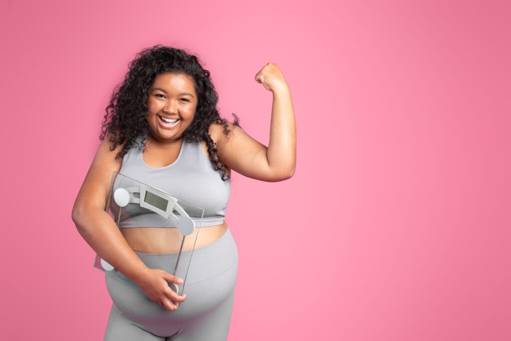 Positive black obese woman in sportswear holding scales and showing muscles biceps, standing on pink studio background, free space. Fitness, body care, strength training and weight loss