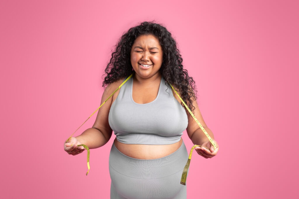 Unhappy sad black plus size woman in sportswear holding measuring tape on neck and crying isolated on pink background. Proper nutrition, healthy lifestyle and motivation concept