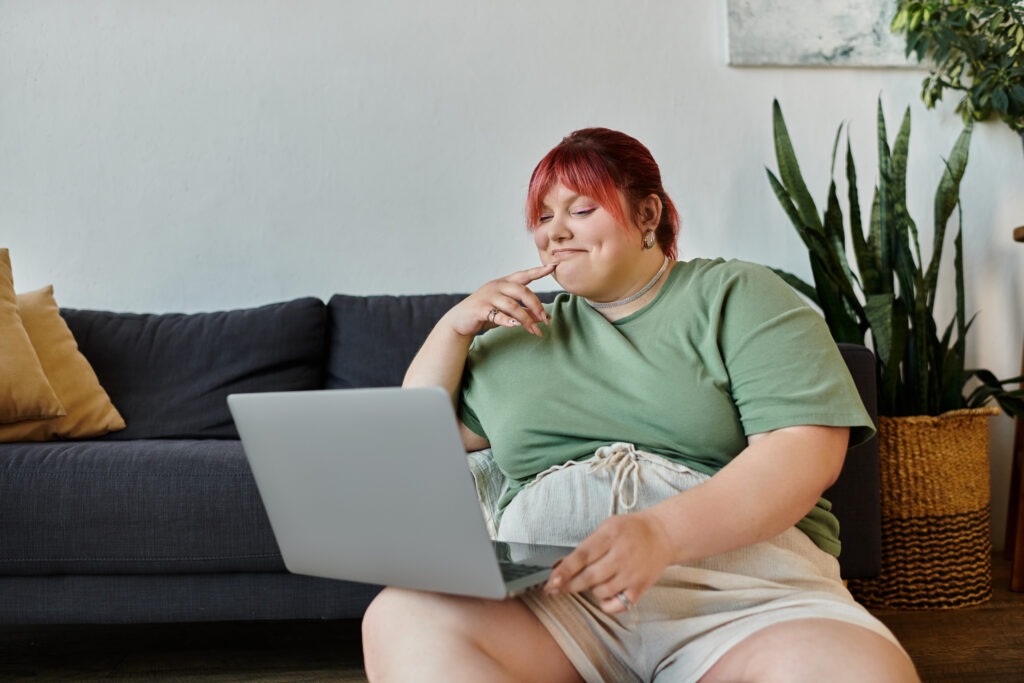 A plus size woman sits on a couch, thoughtfully looking at a laptop.