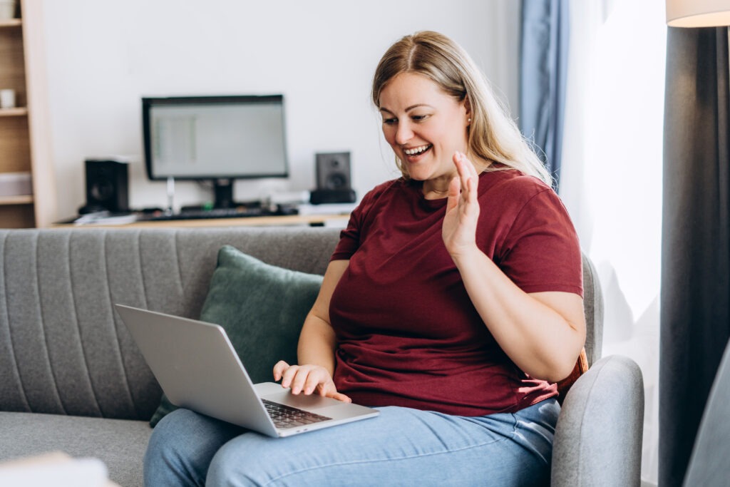 Smiling beautiful plus size woman using laptop, looking at screen, waving hand, speaking online at home