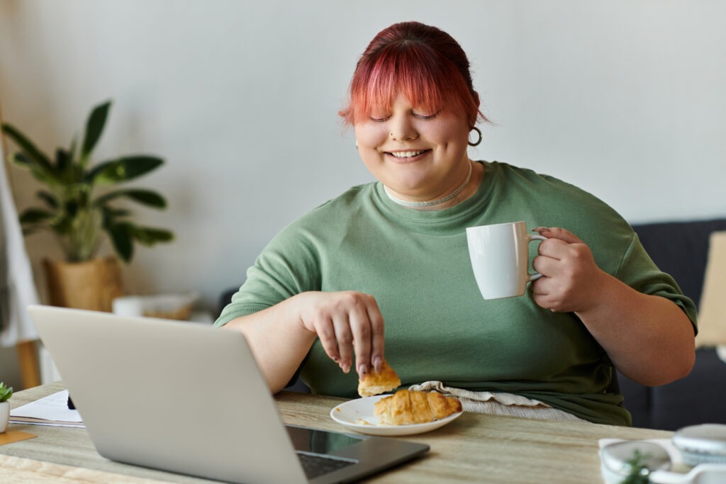 A plus-size woman smiles as she enjoys coffee and pastry while working on a laptop.