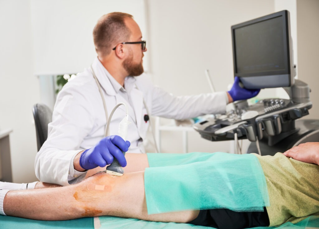 Doctor making ultrasound diagnostics, patient lying on couch. Focus on hand. Male specialist wearing uniform and glasses, looking at screen. Concept of medicine and healthcare.