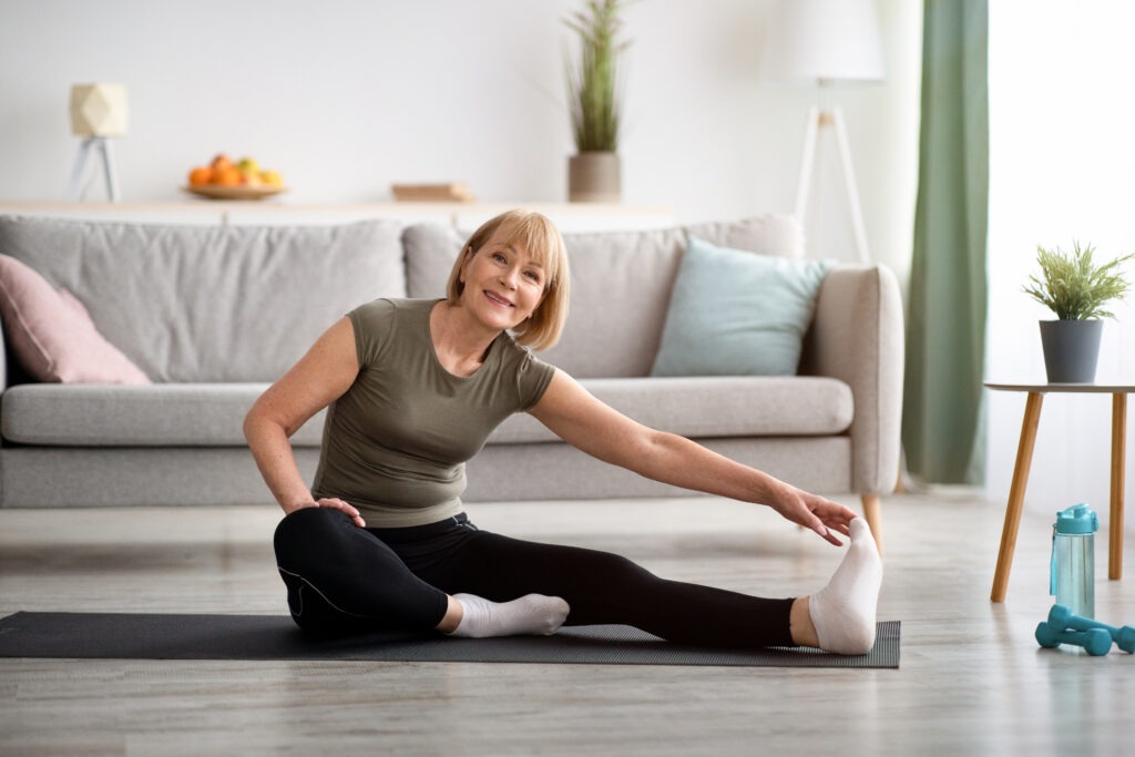 Domestic workout. Cheerful senior lady stretching her leg on sports mat in living room, empty space. Flexible mature woman doing exercises, leading healthy lifestyle during covid lockdown